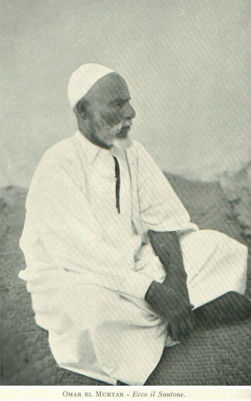 A black and white photo of 'Umar al-Mukhtar. He is wearing white clothes, a white cap, has a well-trimmed beard, and is sitting cross-legged while looking to the right.